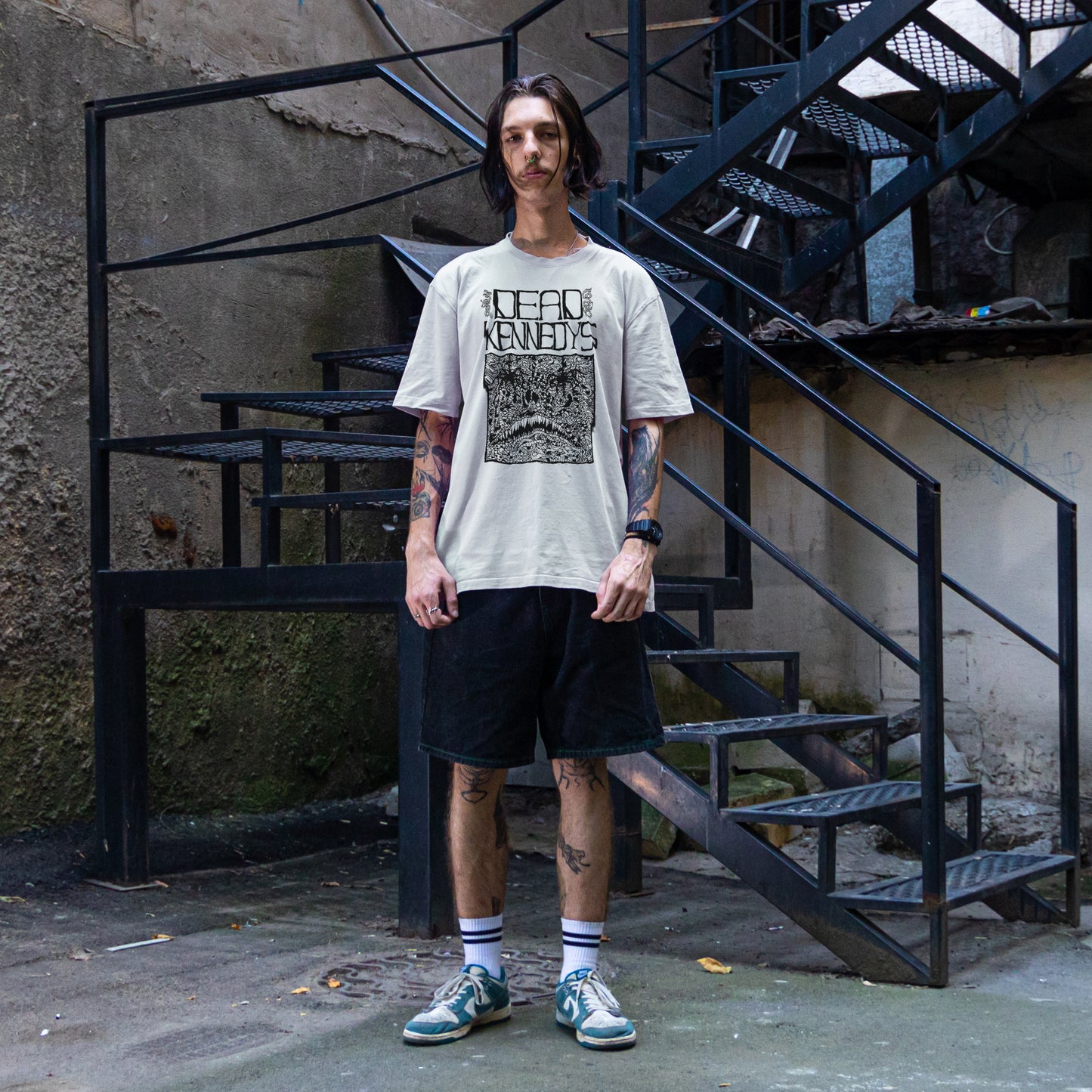 Male model wearing a Dead Kennedys band t-shirt with black shorts and teal sneakers in an urban metal stairwell